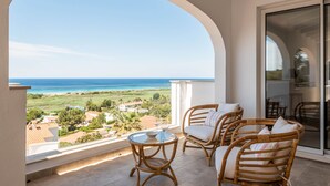 Un balcón con mobiliario de mimbre, una mesa de vidrio y vistas al mar y al paisaje.