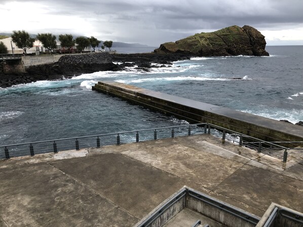 A coastal walkway with a stone pier extending into the sea, a rocky outcrop, and a view of the ocean.