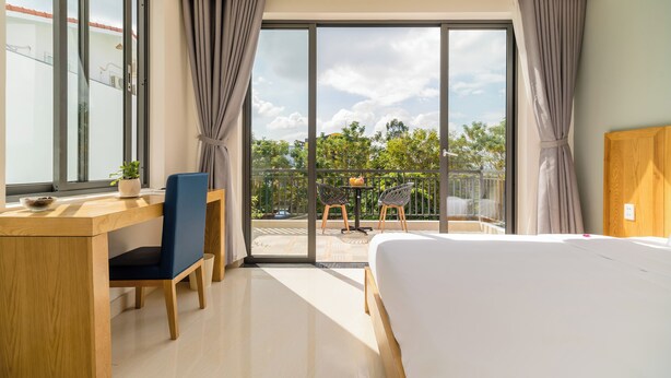 A hotel room with a large bed, a desk with a chair, a balcony with a table and chairs, and a view of trees and buildings.