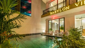 An indoor swimming pool with a glass wall, surrounded by plants and wicker chairs.