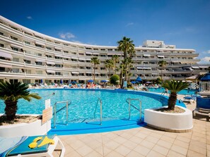 Una piscina amplia rodeada de sillones de descanso y palmeras, con un edificio de hotel de varios pisos al fondo.