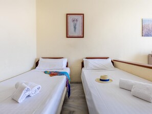 A hotel room with two single beds, white linens, a colorful blanket, a rolled towel, a straw hat, and a framed picture on the wall.