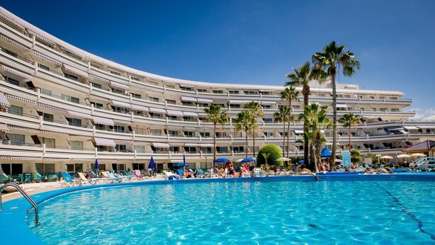 Un hotel grande con múltiples balcones y un área de piscina con tumbonas y palmeras.