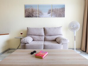 A living room with a beige sofa, a small round table with a wine glass, a book, and a standing fan.