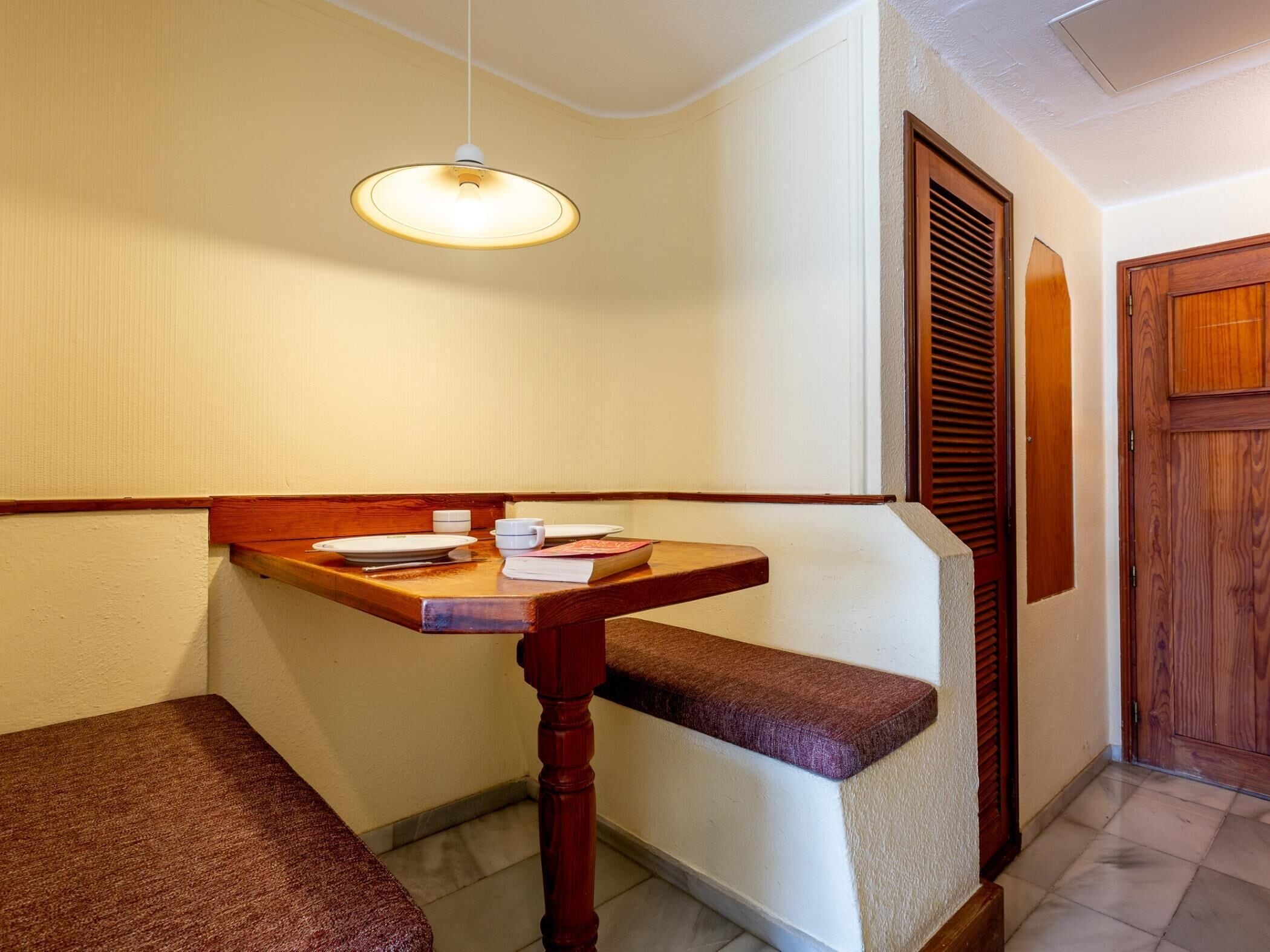 A corner room with a wooden bench, a table with a white plate, a cup, and a book.