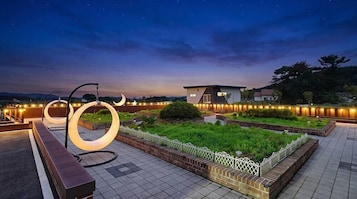 Um jardim no terraço com luminárias circulares, uma cerca branca e um edifício ao fundo.