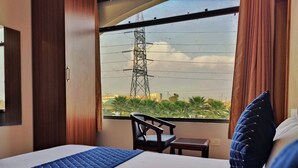 A hotel room with a bed, a chair, a small table, and a window with a view of palm trees and a power line tower. - Hotel Park Hills (Mohali)