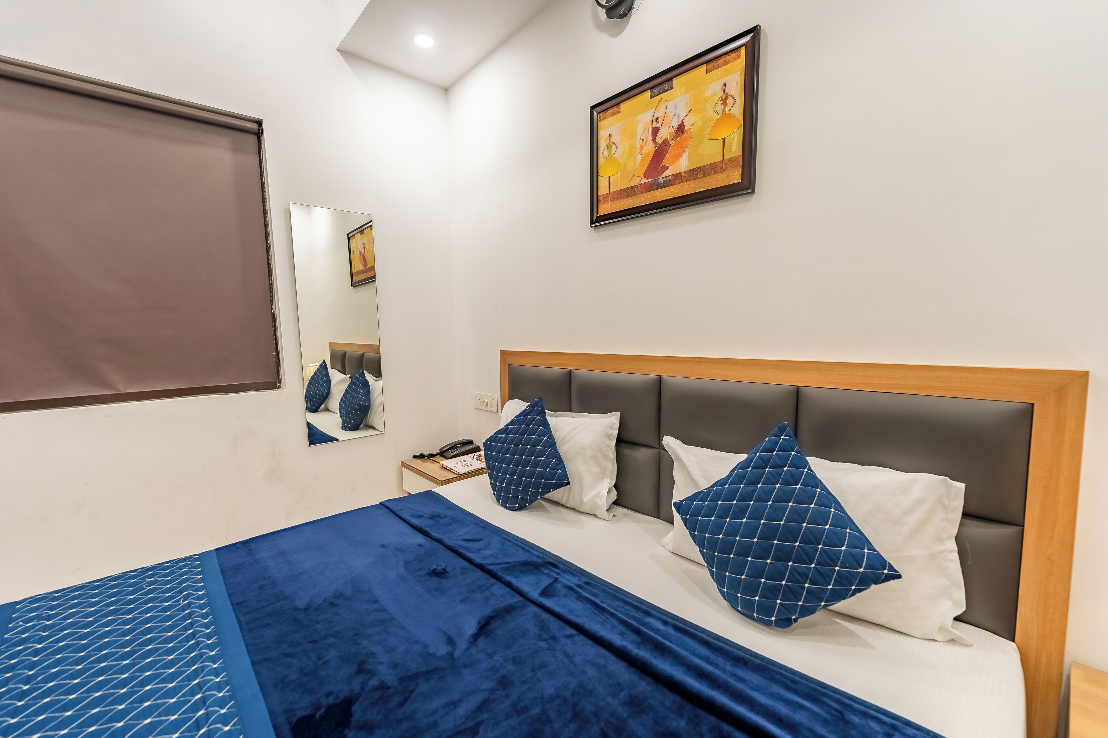A modern hotel room with a large bed, a headboard, a nightstand, a mirror, and a painting on the wall.