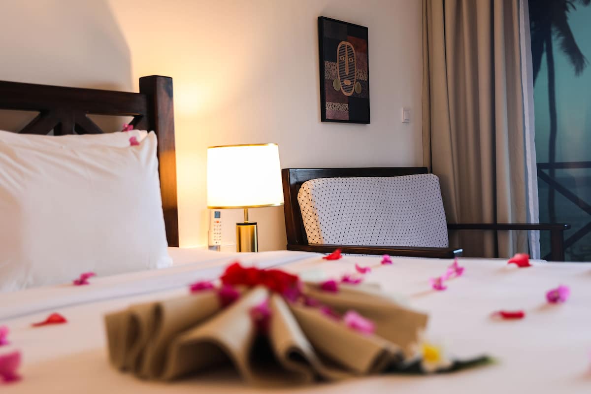 A hotel room with a neatly made bed, a bedside lamp, a framed artwork on the wall, and a view through a window with curtains.