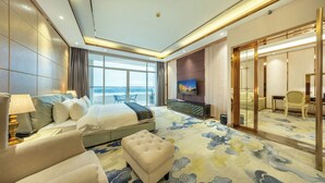 Une chambre d’hôtel moderne dotée d’un grand lit, d’un coin salon avec un canapé et un pouf, d’une télévision et offrant une vue sur la mer grâce à de grandes fenêtres.