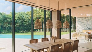 A modern dining area with a large table and chairs, large windows, and a view of a swimming pool and greenery. - Villa El Cielo Ishigaki (Ishigaki)
