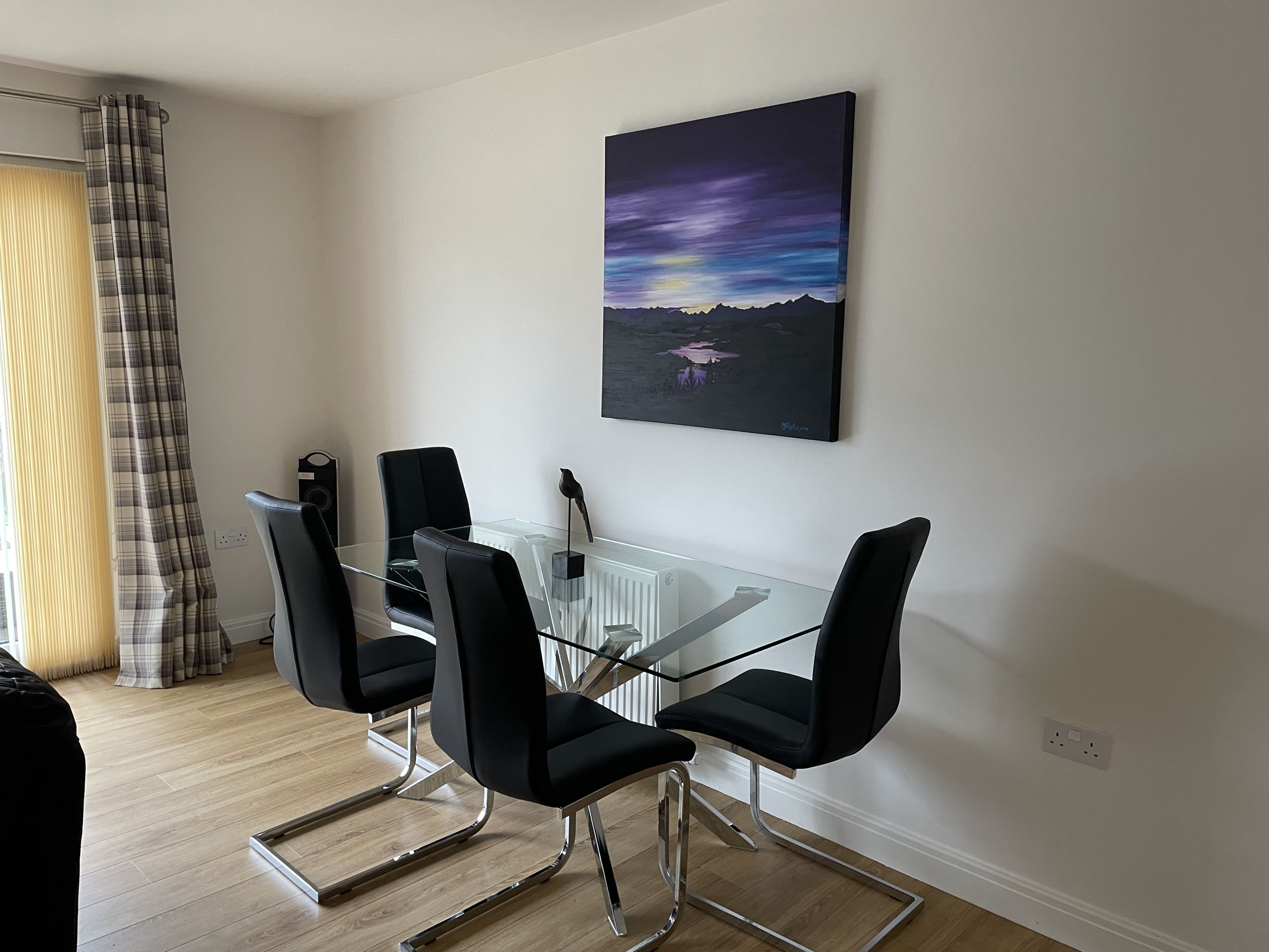 A modern dining area with a glass table and black chairs, a painting of a landscape on the wall, and a plaid curtain.