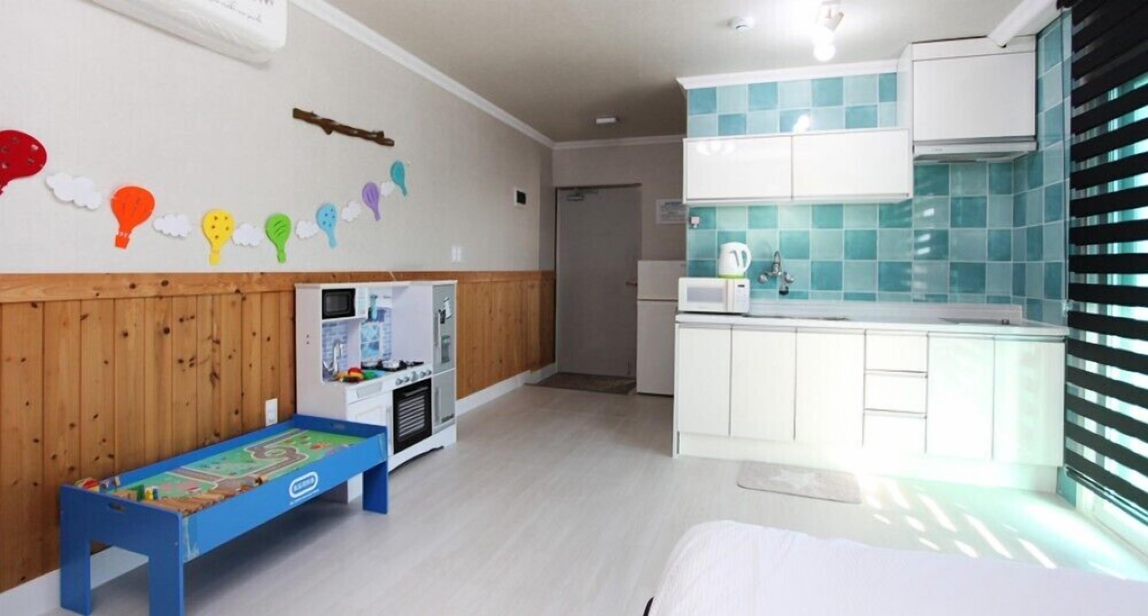 A room with a wooden wall, a kitchenette with white cabinets, a blue play table, and a bed with white bedding.