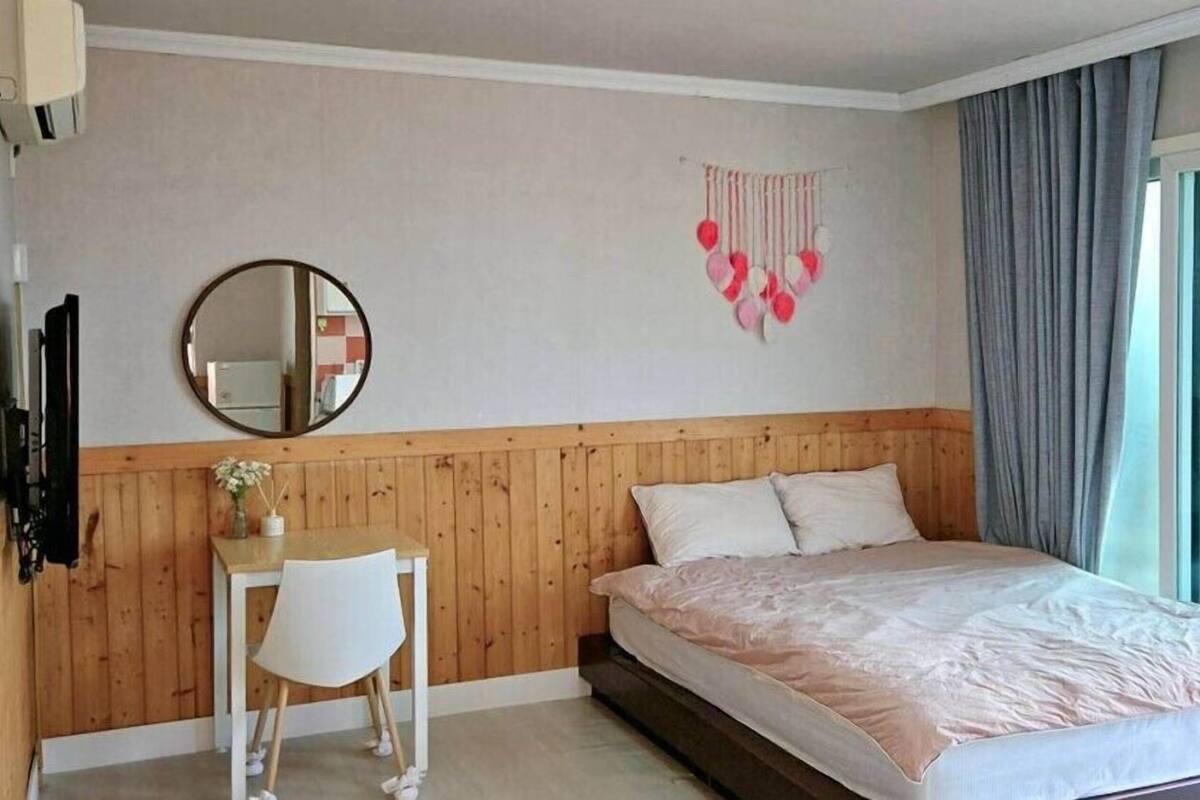 A bedroom with a wooden headboard, a bed with white bedding, a round mirror, a small desk with a chair, and a wall decoration.