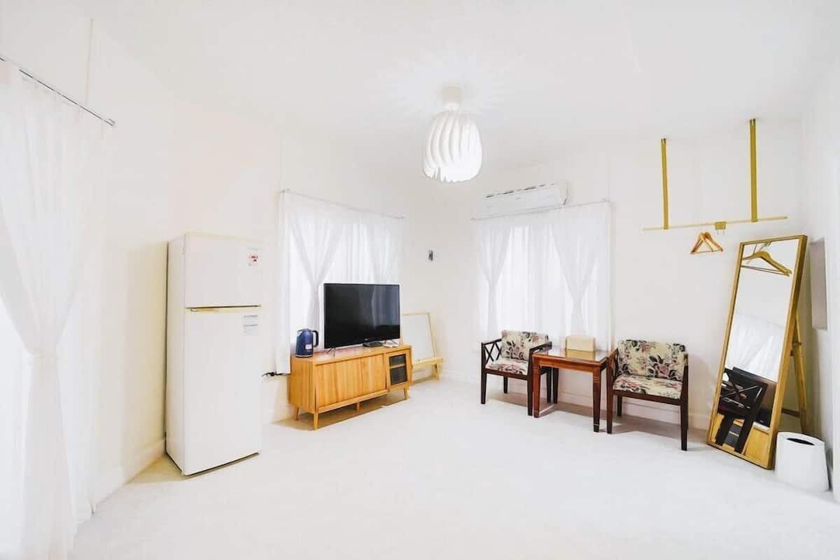 Un salon minimaliste avec un réfrigérateur blanc, un meuble TV en bois, un téléviseur à écran plat, deux fauteuils à motifs, une petite table et un grand miroir.