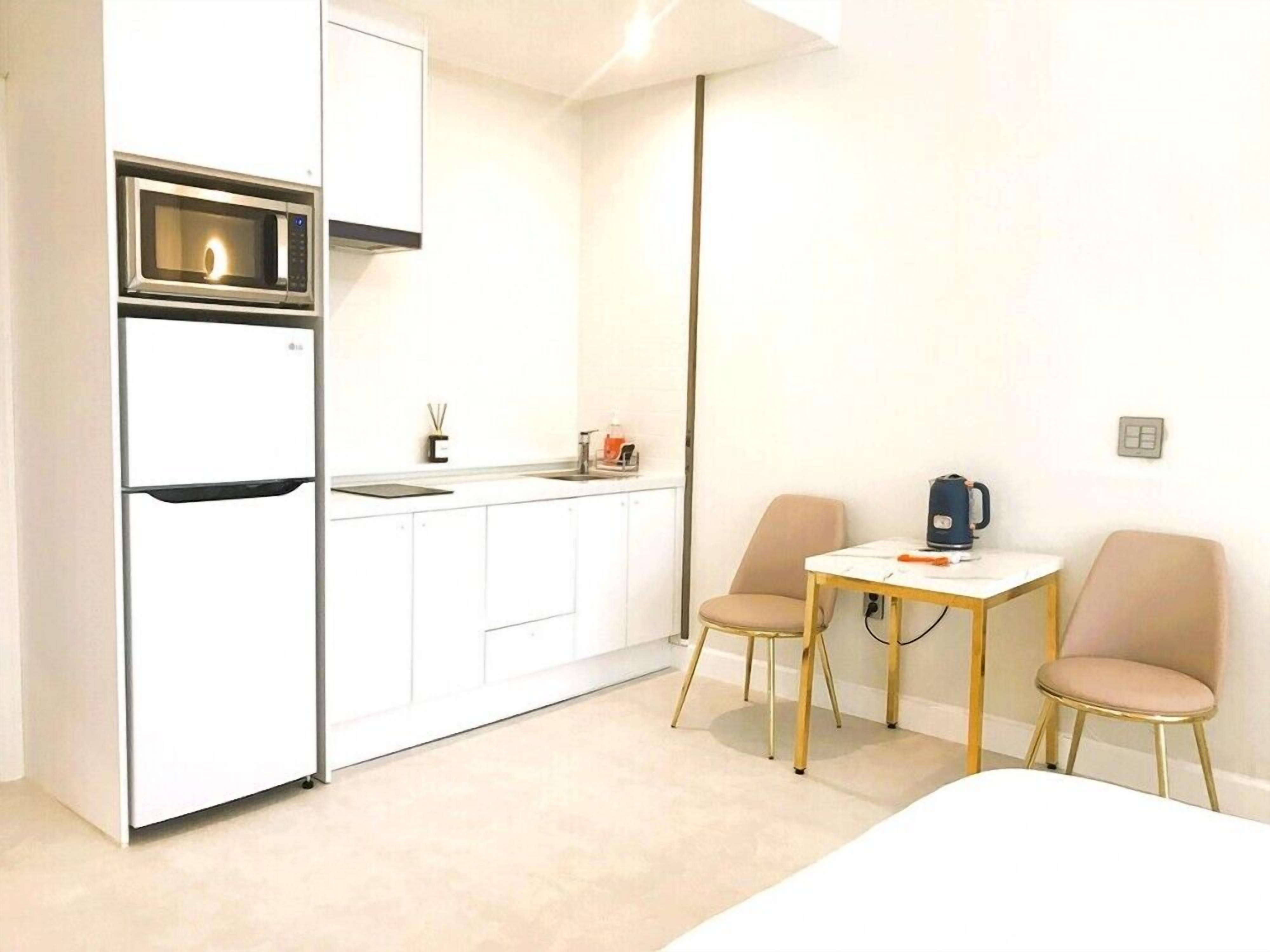 A compact kitchen with a microwave, refrigerator, and sink. There is a small table with two chairs and a kettle.