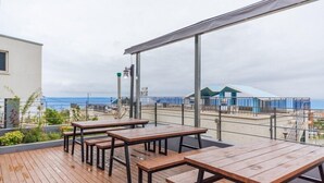 A wooden deck with tables and benches overlooking a beach and a building with a blue roof. - Heart Blue Road Town Pension (Yeongdeok)
