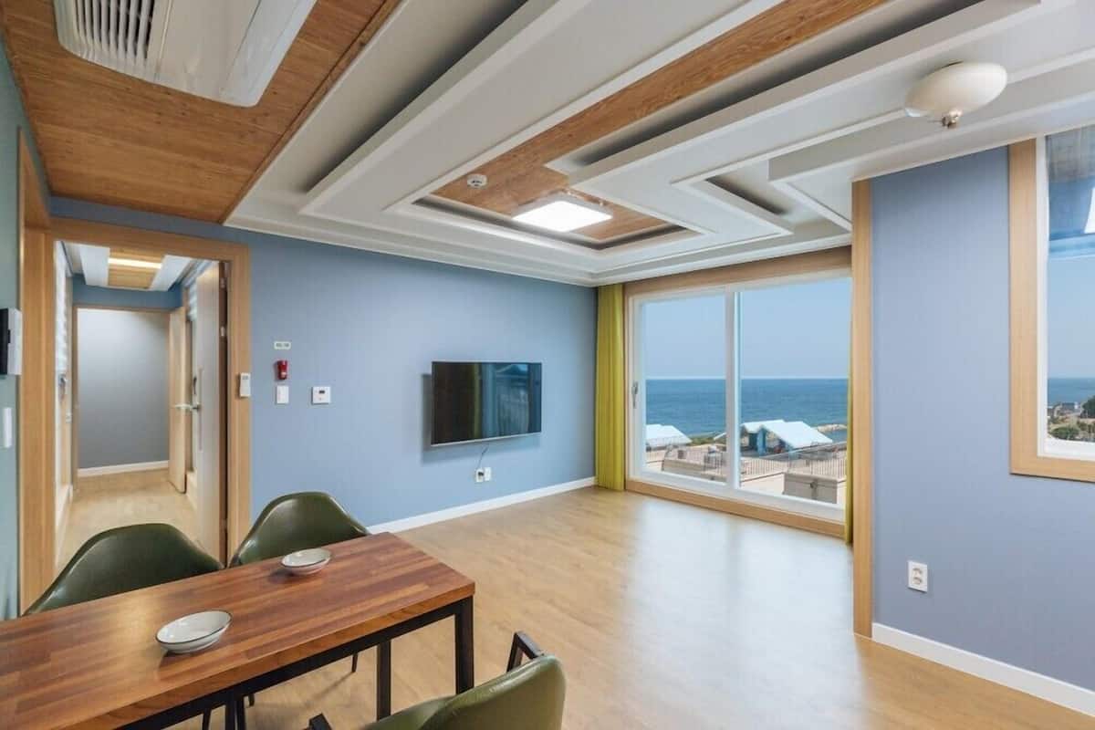 Una habitación con techo de madera, televisor de pantalla plana, mesa de comedor con sillas y un amplio ventanal con vista al mar.