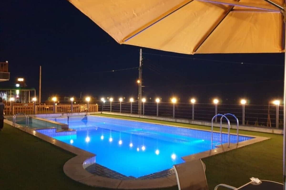 Una piscina nocturna con bordes iluminados, rodeada por una valla y una pérgola.