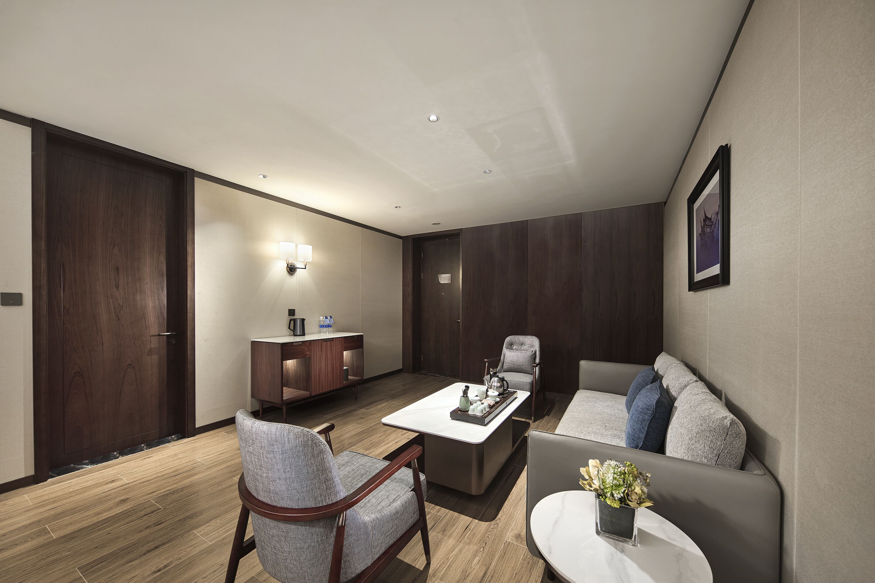 A modern hotel room with a wooden floor, a sofa, a chair, a coffee table, and a small side table with a vase.