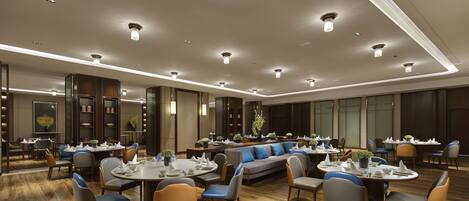 A spacious hotel dining area with multiple round tables, comfortable seating, and modern lighting fixtures.
