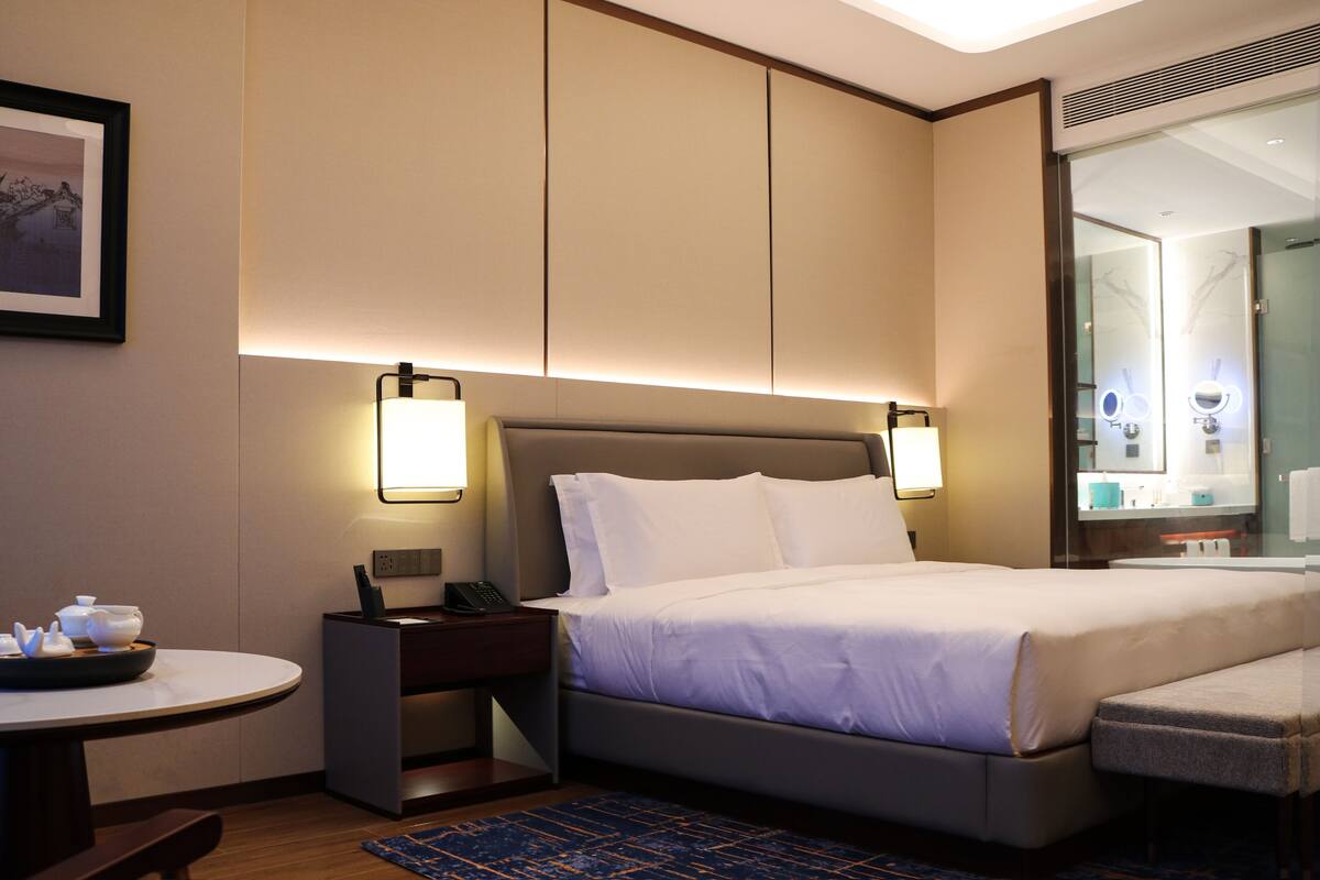 Una habitación de hotel con cama, mesita de noche, lámpara y una pequeña mesa redonda con tetera.
