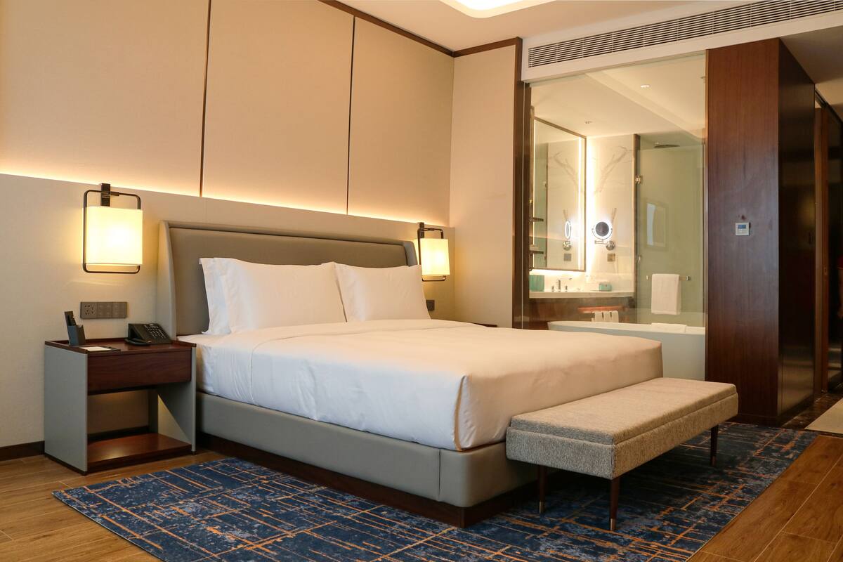 Habitación de hotel con cama, mesitas de noche, un banco, baño con espejo y ducha.