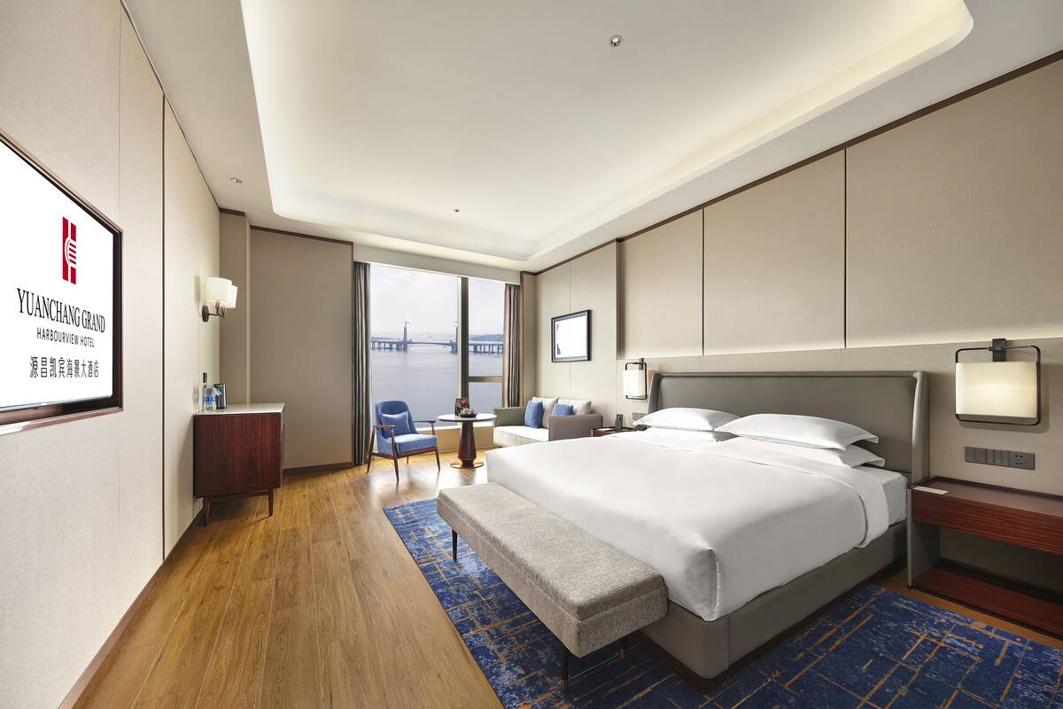Habitación de hotel con una cama grande, un escritorio, una silla, un banco y vista a un puente.