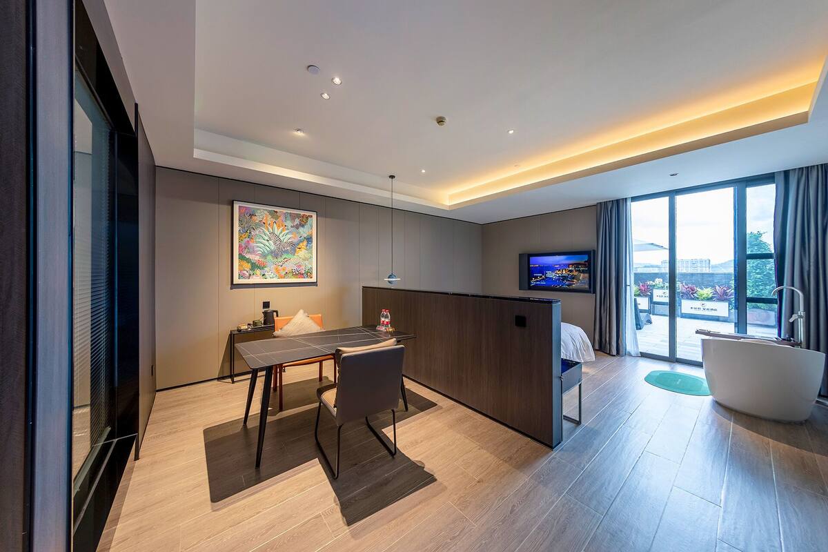 A modern hotel room with a dining area, a large window with a view, a flat-screen TV, and a bathtub.