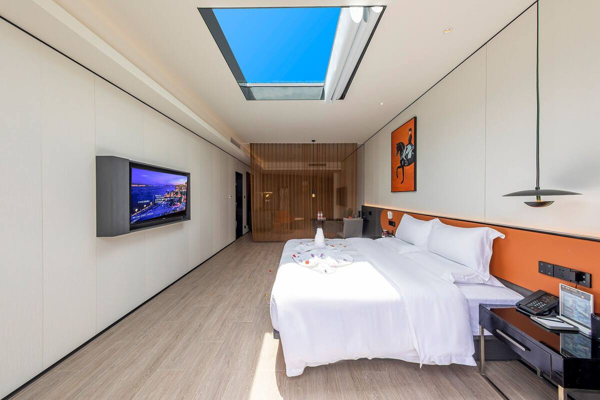 A modern hotel room with a large bed, a desk, a flat-screen TV, and a skylight.