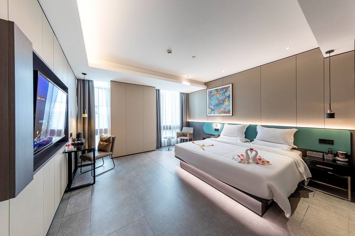 A modern hotel room with a large bed, a desk, a chair, a TV, and a painting on the wall.