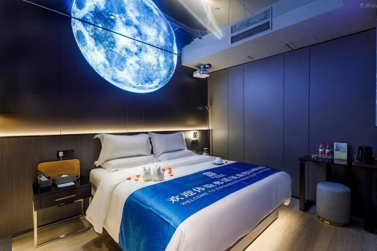 A modern hotel room with a large bed, a nightstand, a small table, and a wall-mounted projector displaying a moon.