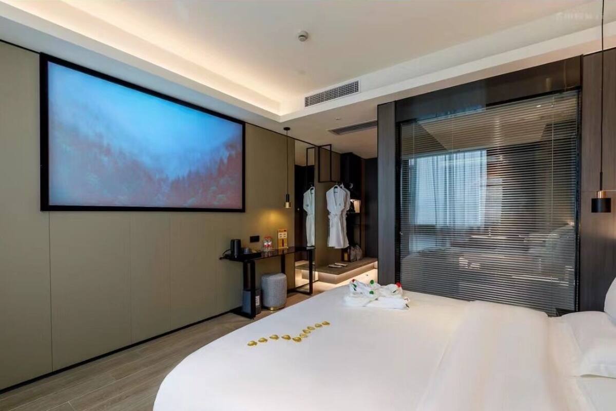 A modern hotel room with a large bed, a flat-screen TV, a mirror, and a window with blinds.