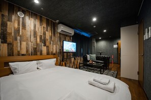 A modern hotel room with a wooden wall panel, a large bed with white linens, a flat-screen TV, a sofa, and a small table with a remote control. - SILK COURT 202 (Chitose)