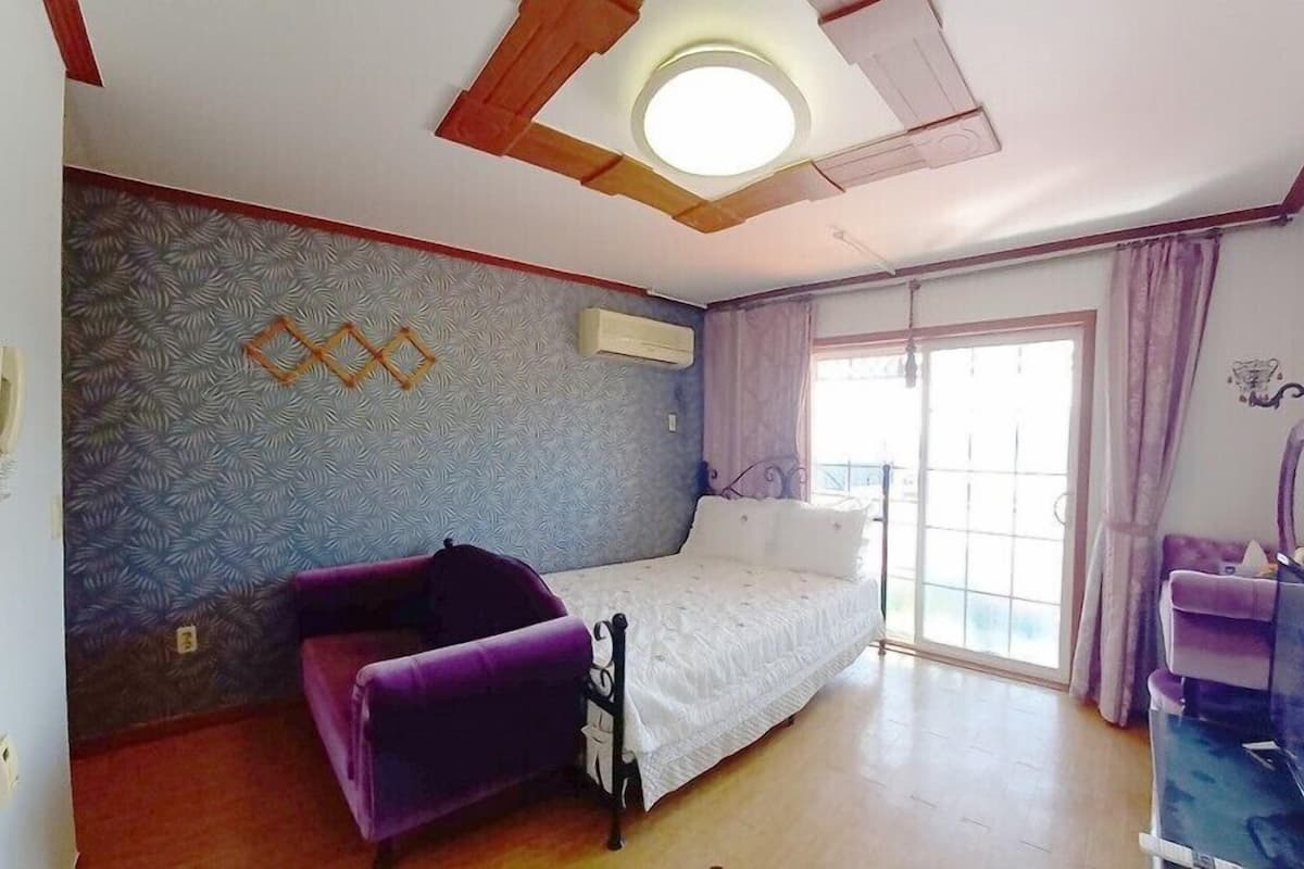 A room with a bed, a purple chair, a TV, and a window with curtains.