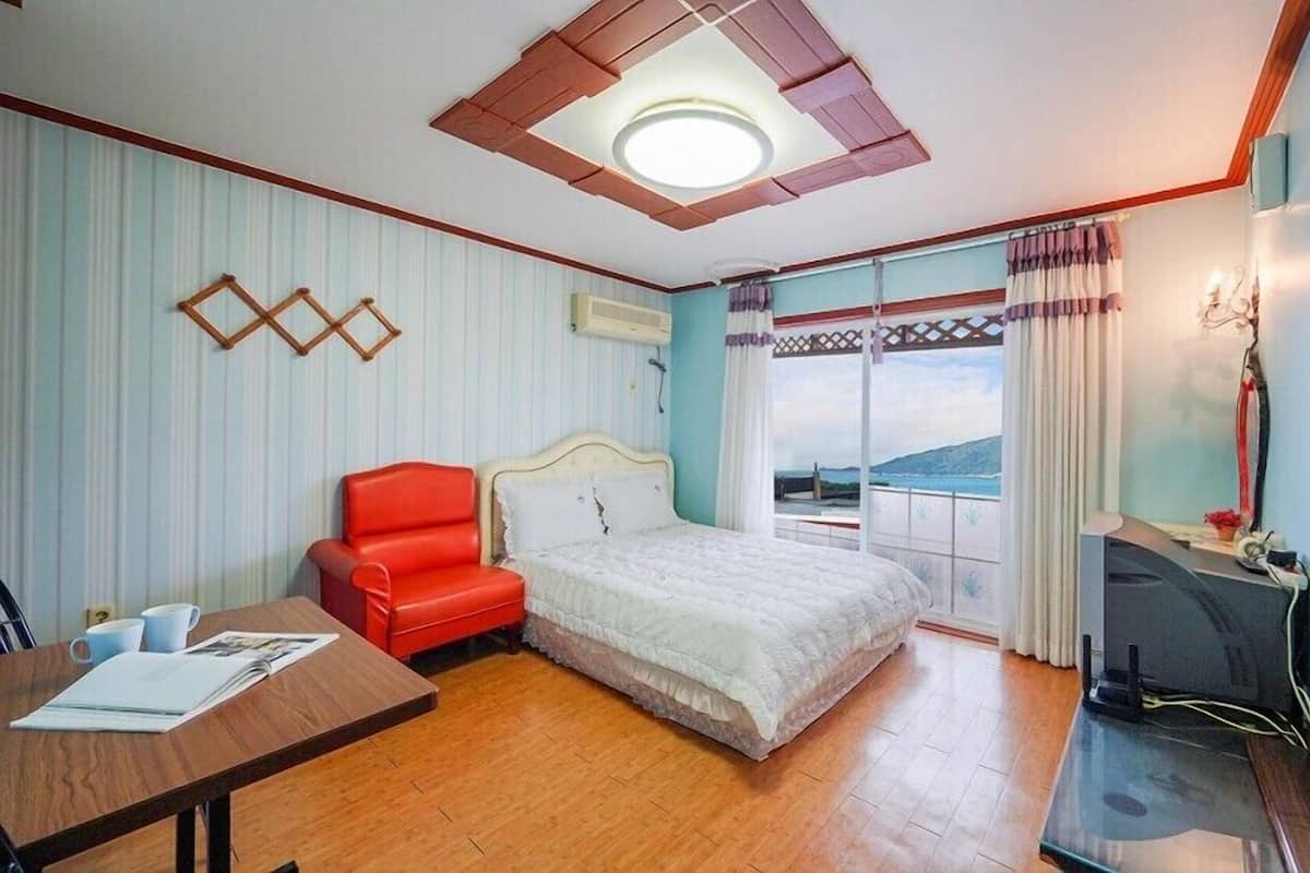 A hotel room with a bed, a red chair, a desk, a TV, and a balcony with a view.