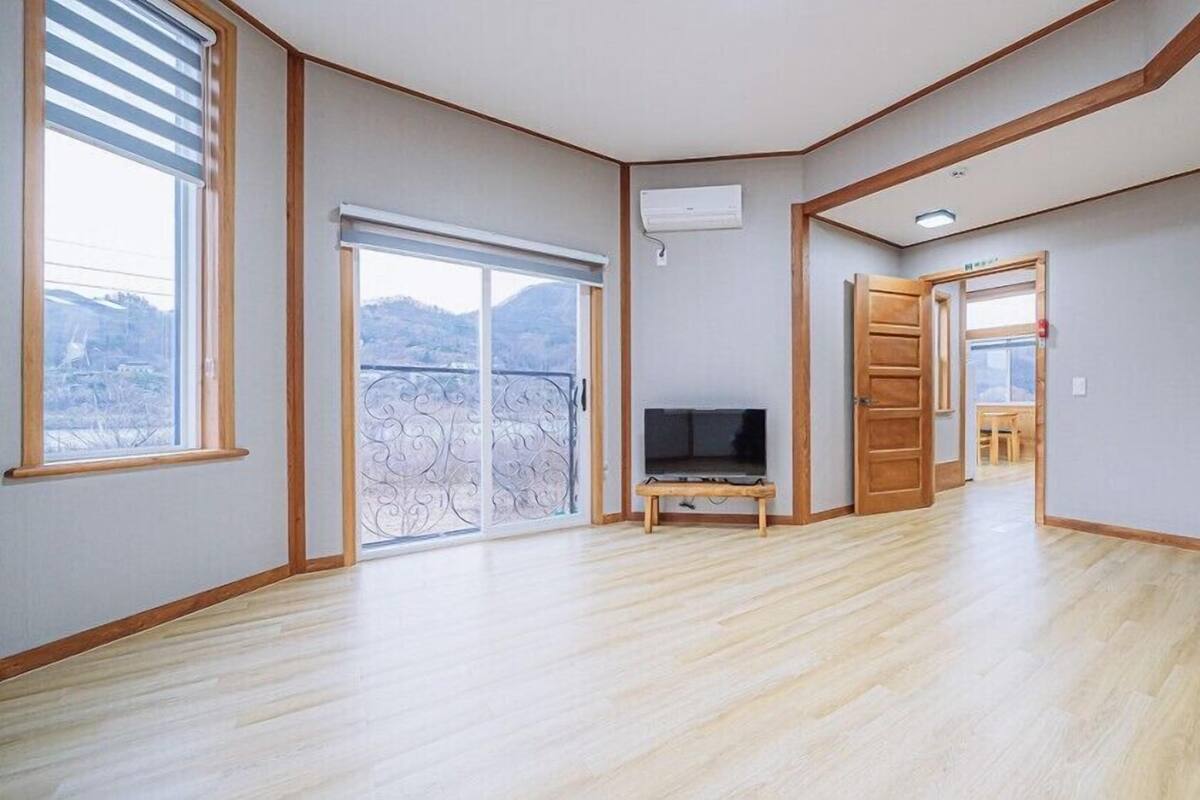 A spacious room with a large sliding glass door leading to a balcony, a flat-screen TV on a wooden stand, and a wooden door leading to another room.