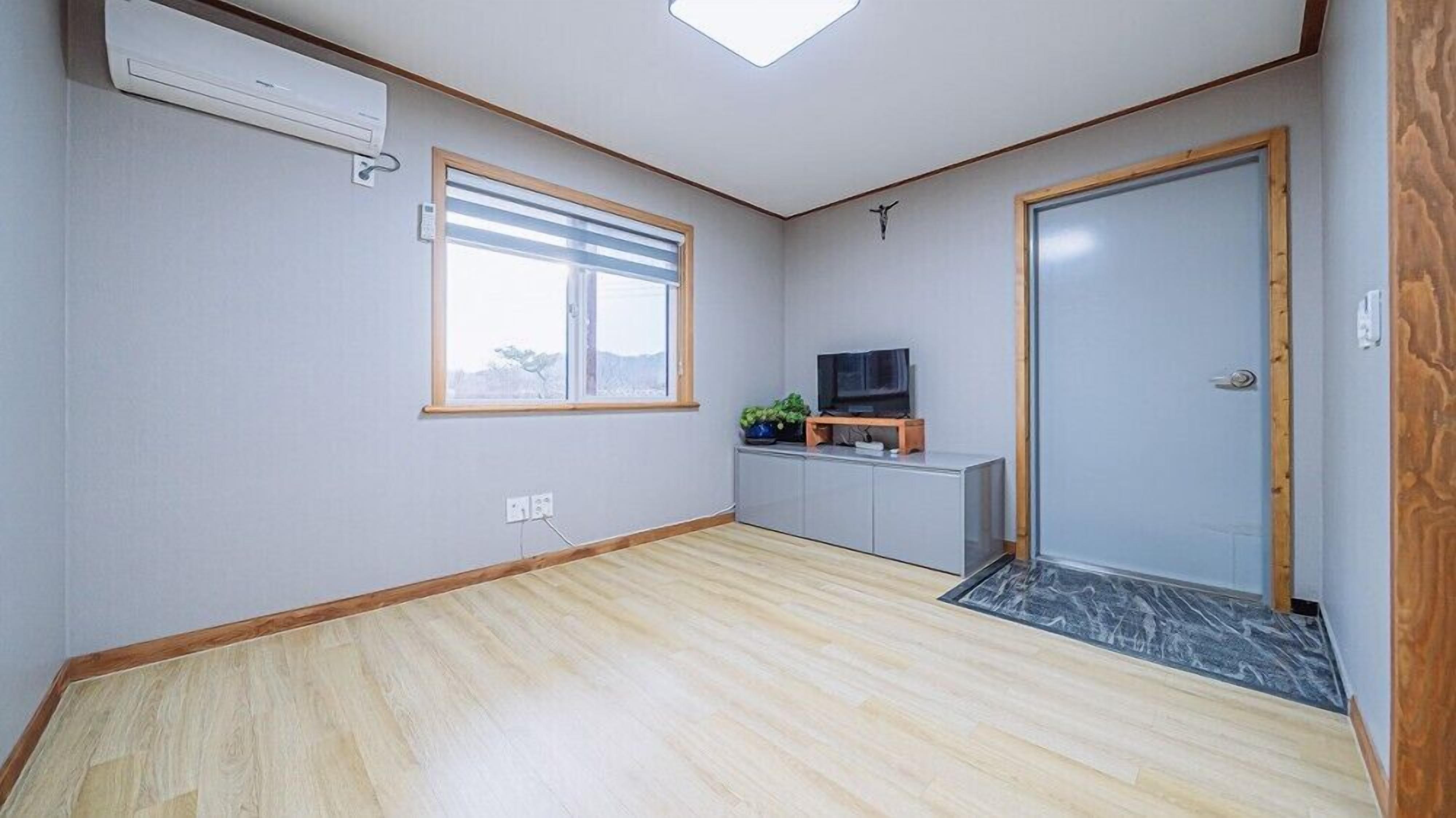 A room with a wooden floor, a window with blinds, a television on a stand, a door, and a small plant.