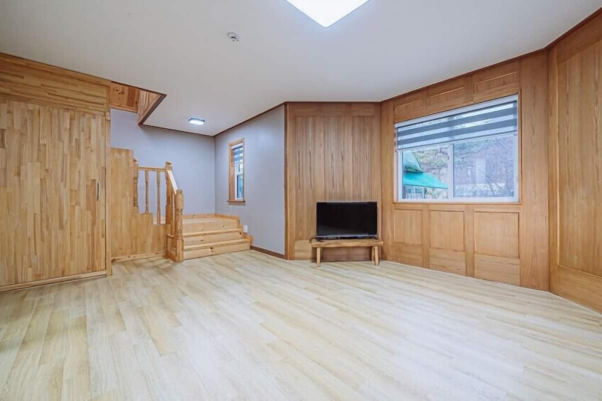 A spacious room with a wooden floor, a built-in TV, a staircase, and a large window with blinds.