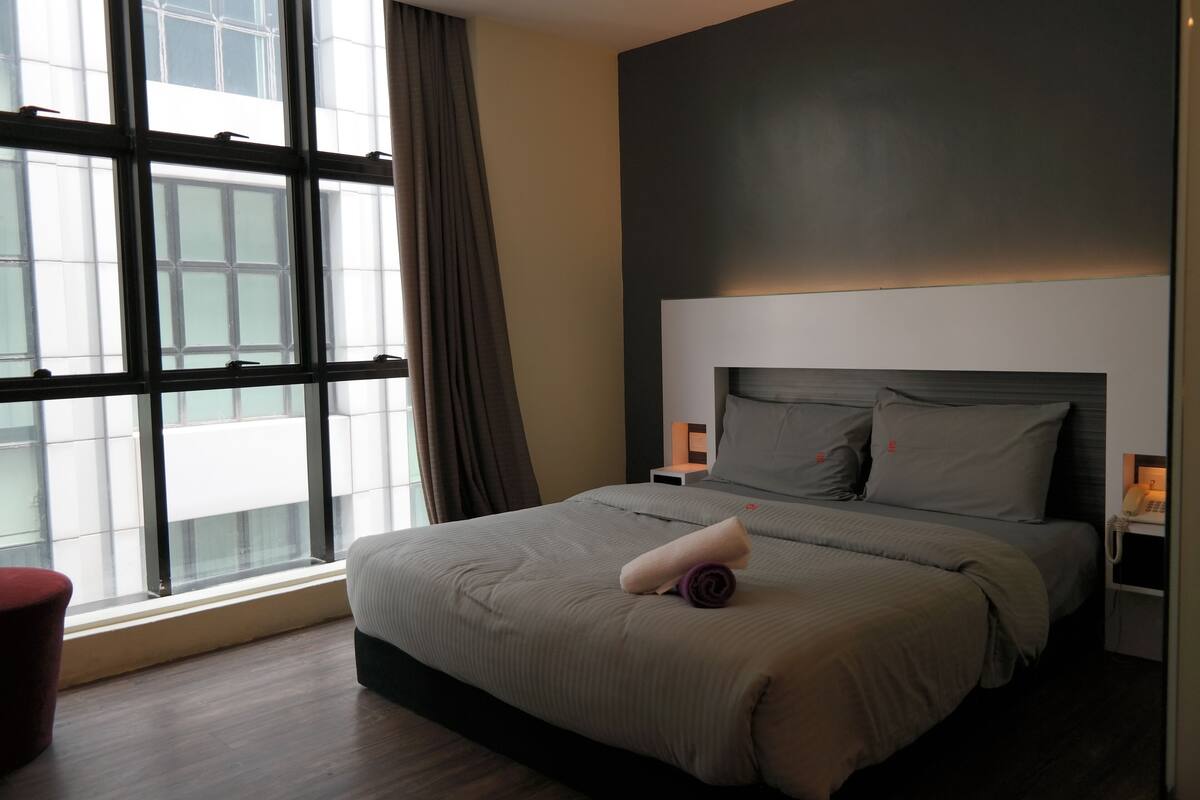 Habitación de hotel moderna con una cama grande, una mesita de noche con lámpara y vistas a través de ventanales del suelo al techo.