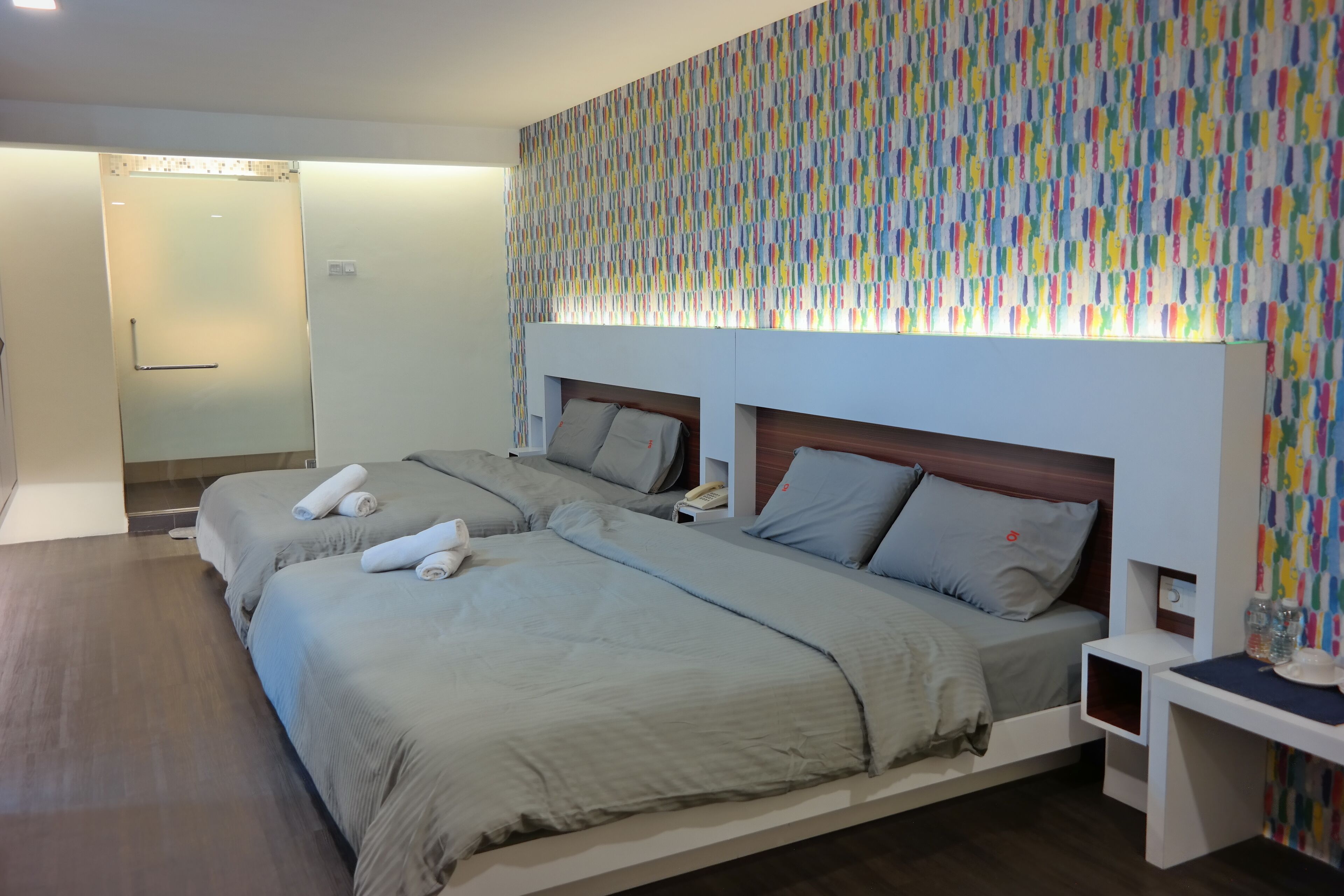 A hotel room with two beds, a wooden floor, a colorful wall, and a small bedside table.