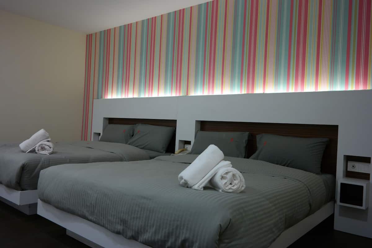 Dos camas individuales con ropa de cama gris y cabeceros blancos, una pared a rayas y una mesita de noche pequeña con una lámpara.
