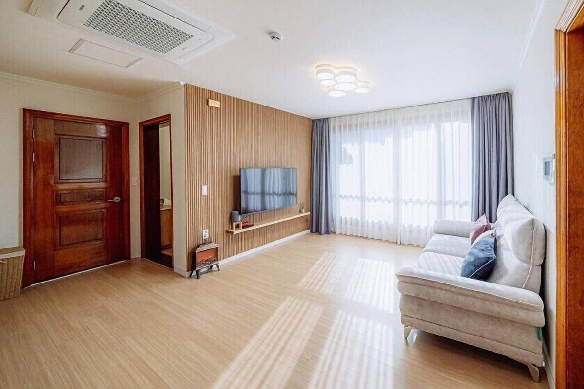 Ein geräumiges Wohnzimmer mit Holzboden, einer Couch, einem an der Wand montierten Fernseher und einem großen Fenster mit Vorhängen.