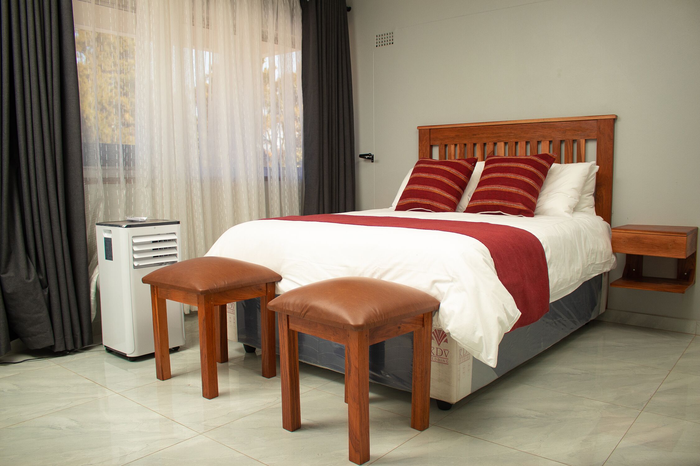 Una cama bien tendida con cabecera de madera, una mesita de noche y dos taburetes de madera.