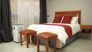 Una cama bien tendida con cabecera de madera, una mesita de noche y dos taburetes de madera.