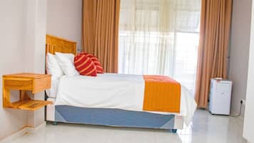 Una cama bien tendida con cabecera de madera, una mesita de noche con cajón y un pequeño electrodoméstico blanco, probablemente un calentador, en una habitación luminosa con ventanas grandes y cortinas.