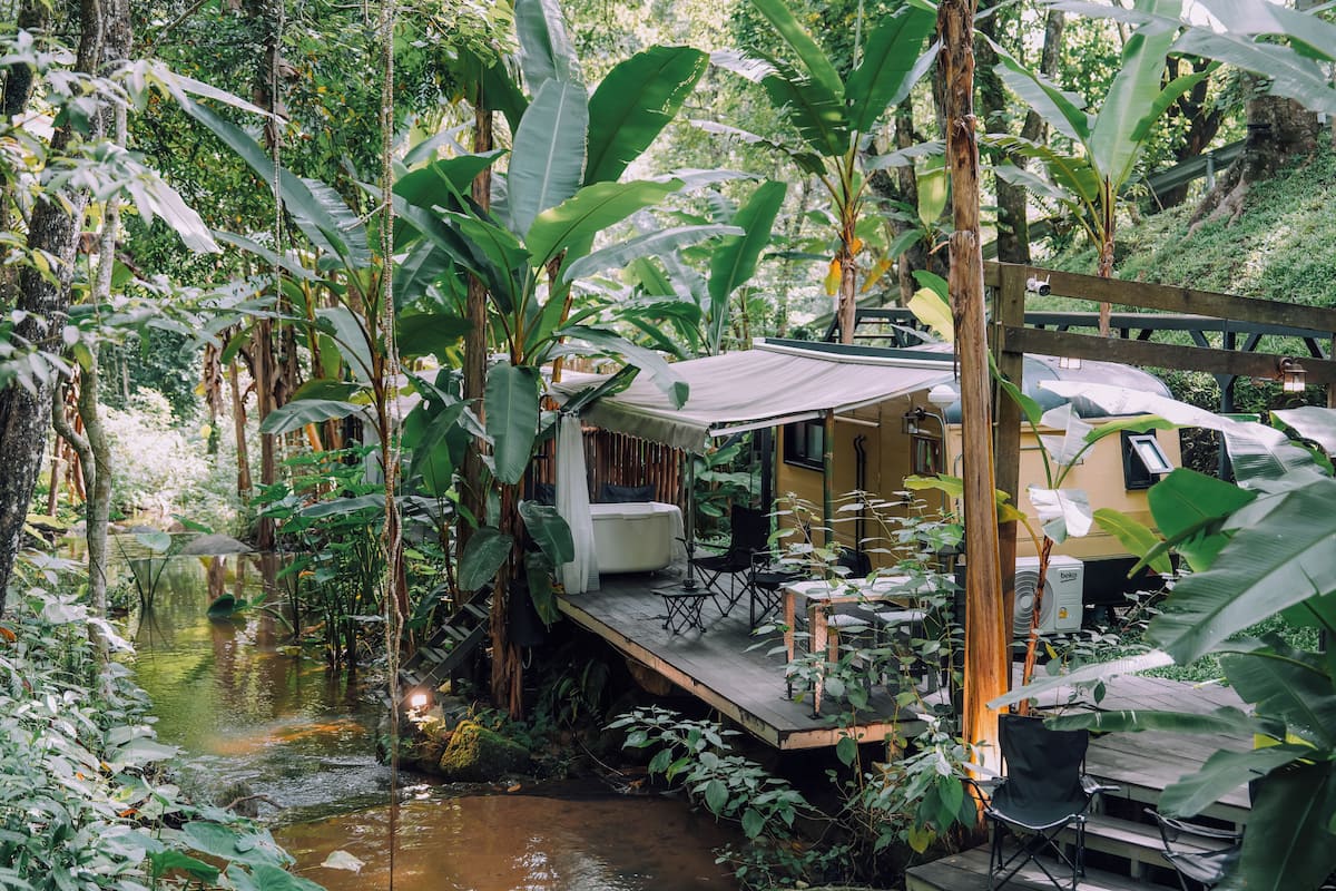 Terraza de madera con mobiliario exterior, rodeada de exuberante vegetación tropical y una pequeña extensión de agua.