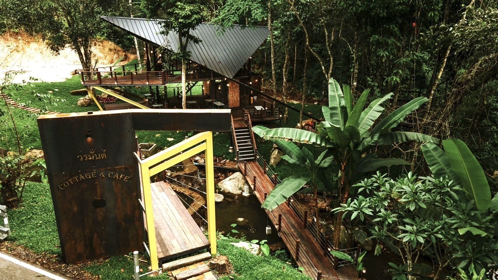 Une construction rustique en bois, portant l’inscription « COTTAGE & CAFE », avec une terrasse en bois et un petit étang, le tout entouré d’une végétation luxuriante.