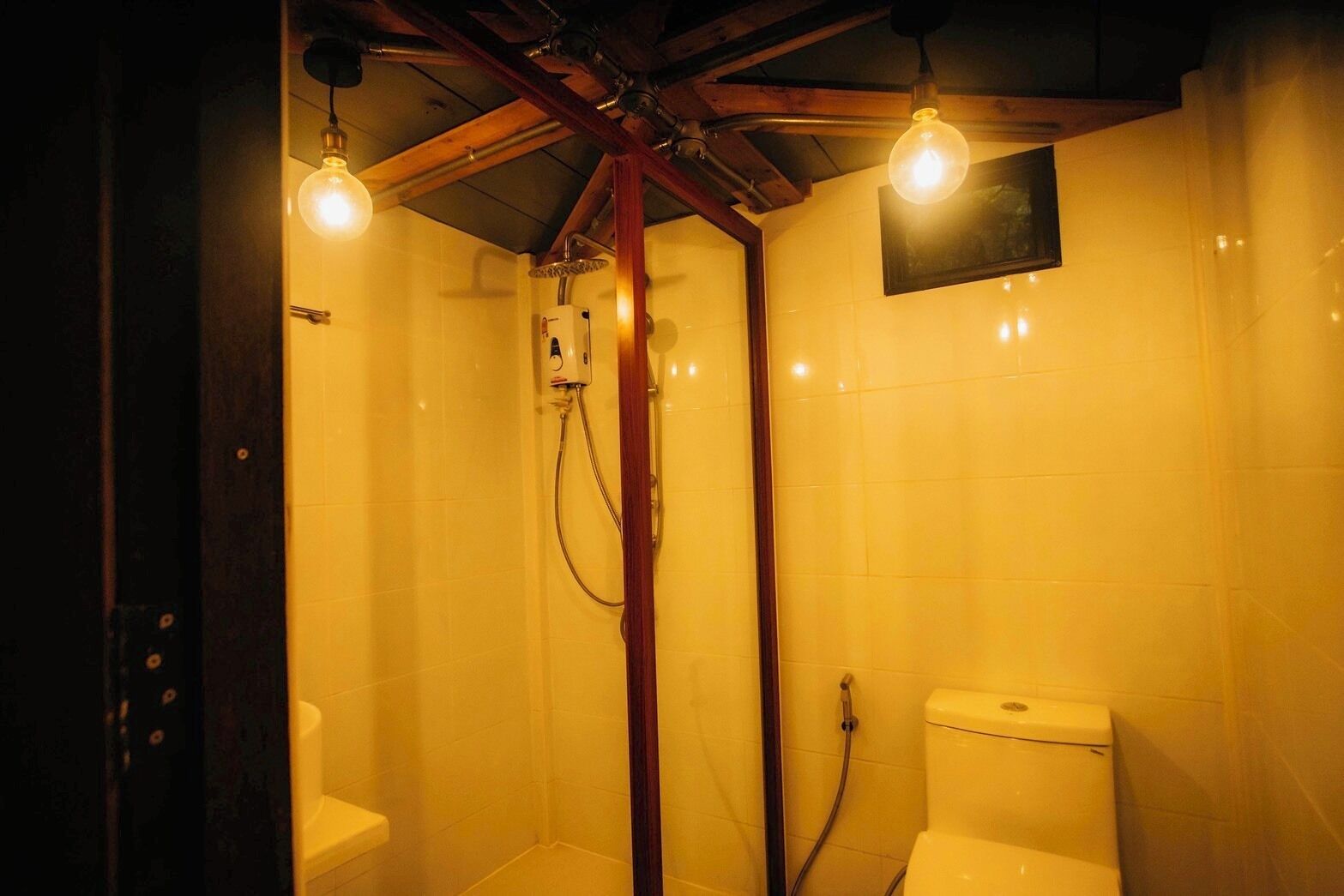 A small, compact bathroom with a shower and toilet, wooden beams on the ceiling, and two exposed light bulbs.