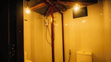 A small, compact bathroom with a shower and toilet, wooden beams on the ceiling, and two exposed light bulbs.